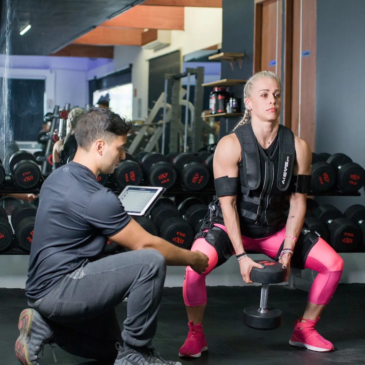 Antrenör EMS cihazını tabletten ayarlarken EMS kıyafetli bir kadın dambılla squat yapıyor